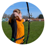 Carris drawing a recurve bow while practising archery on an outdoor grass field with targets in the background.