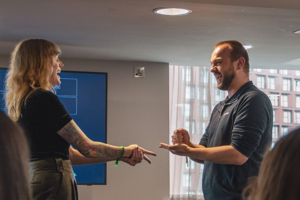 Two GuyKat colleagues laughing while playing rock, paper, scissors during a team-building workshop in a modern office.